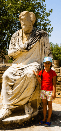 Girl with red tshirt stands next to a bog Greek statue in Crete